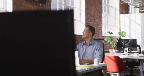 Young professional typing on laptop in bright industrial open-plan office with loft windows
