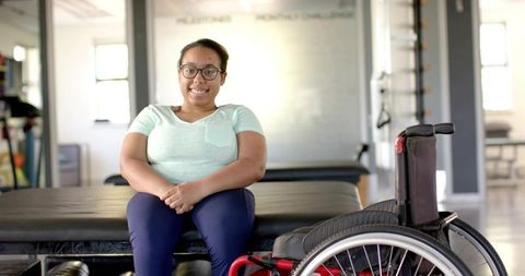 Smiling woman sitting on therapy table wearing mint t-shirt and navy leggings next to wheelchair