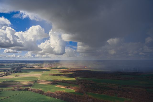 Dramatic sky with large clouds casting shadows over vast green farmlands with a rainstorm approaching in the distance. This aerial view offers a unique perspective of the countryside, ideal for weather-related projects, agriculture content, or rural scenery presentations.