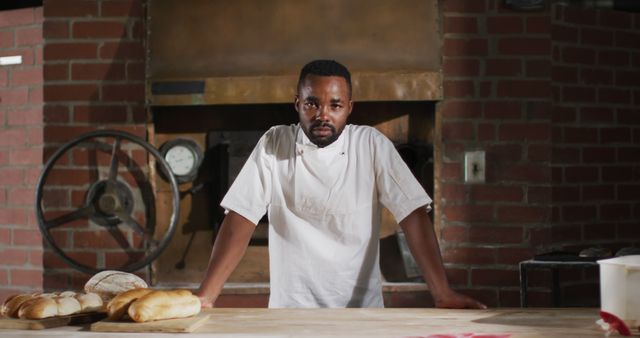 Shows a confident baker standing in a rustic brick bakery with fresh bread displayed on the table. Ideal for use in promoting artisanal baking, bakery businesses, culinary training programs, chef profiles, and food industry campaigns.