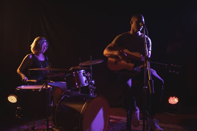 Male singer playing guitar and female drummer performing on stage in a dimly lit nightclub. Ideal for use in articles about live music, nightlife entertainment, band performances, and concert promotions.