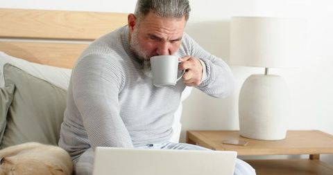 Mature man sipping coffee while working on laptop in cozy bedroom with sleeping dog