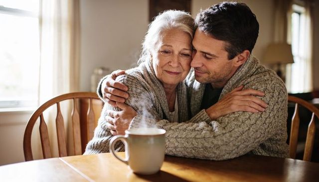 Adult son embracing senior mother at wooden dining table with steaming mug, cozy warmth