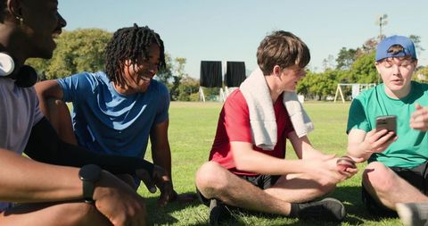 Young athletes relaxing on grass after practice laughing, sharing phone, showing teamwork