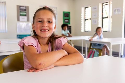 Preteen girl smiling and resting arms on desk in bright classroom with classmates