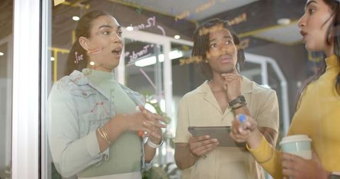Diverse young team brainstorming at glass board with tablet and coffee in modern office