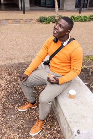 African american man sitting on bench in orange hoodie holding pastry and coffee