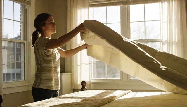 Woman making bed in sunlit bedroom lifting duvet for morning routine and calm interior
