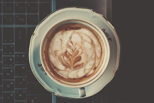Overhead view of a beautifully crafted latte with intricate art in an elegant porcelain cup placed on a laptop keyboard. Ideal for illustrating themes of work, productivity, morning routines, cafe culture, and the coffee break during office hours. Suitable for blogs, websites, social media, and advertisements related to technology, coffee brands, office lifestyles, or freelancers.