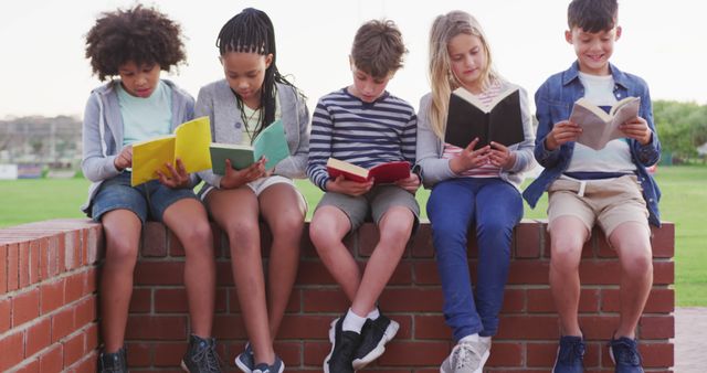 Five diverse children sitting on a brick wall are reading books together outdoors. They appear to be engaged and content, creating a joyful and enriching visual of childhood and education. This image is perfect for educational promotions, campaigns focusing on literacy and learning, library advertisements, or any content related to children's books and reading groups.