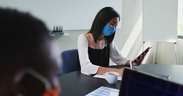 Asian woman wearing face mask using digital tablet while sitting on her desk at modern office. social distancing quarantine lockdown during coronavirus pandemic