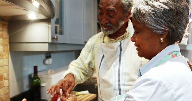 Senior African American couple enjoys cooking together in their kitchen, sharing a joyful moment. They bond over meal preparation, adding spices and ingredients to their dish. This image can be used for lifestyle topics, senior living, family togetherness, cooking blogs, and home kitchen advertisements, illustrating the joy of spending time together at home.