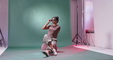 Female model adjusting sunglasses while sitting on pink pouf against green studio backdrop