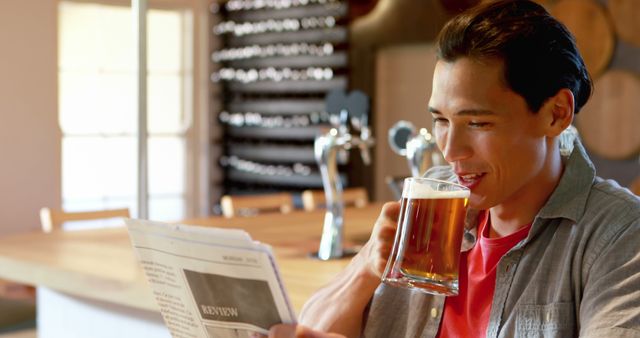 Image depicts a smiling man reading a newspaper while enjoying a glass of beer in a modern pub setting. This can be useful for websites and advertisements related to pubs, leisure activities, and lifestyle content. Ideal for campaigns promoting relaxation, casual dining, and male bonding over drinks.