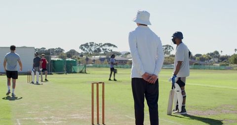 Cricket umpire standing with hands clasped behind back while batsman waiting at crease