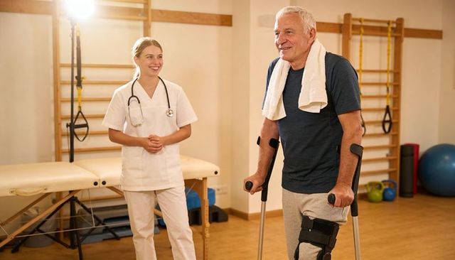 Senior man using forearm crutches and knee brace during physiotherapy with supportive therapist