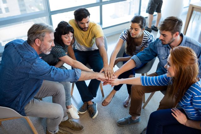 Group of diverse colleagues sitting in a circle, putting their hands together in a gesture of unity and teamwork. Ideal for illustrating concepts of collaboration, team spirit, and workplace cooperation. Suitable for business presentations, team-building workshops, and corporate websites.