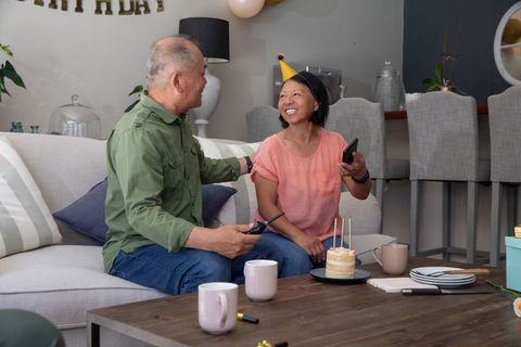 Asian couple smiling and celebrating cozy at-home birthday with small cake and smartphone