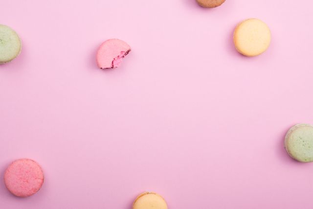 Pastel-colored macarons are spreading across a pink background. One of the macarons, with a bite taken. Perfect for use in bakery promotions, dessert menus, food blogs, or packaging design for confectioneries.