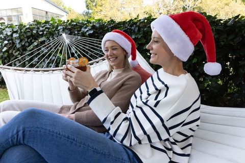 Middle-aged women enjoying backyard holiday cocktails on hammock wearing santa hats