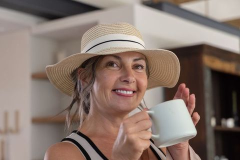 Smiling middle-aged woman holding ceramic mug wearing straw hat in cozy kitchen