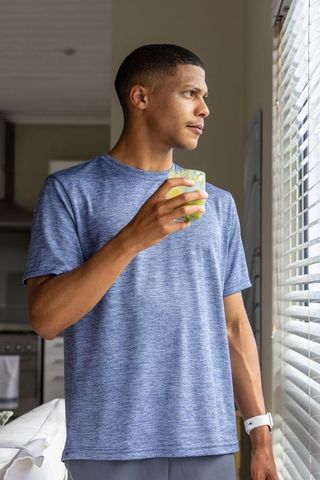 African american man standing at window holding green drink in minimalist kitchen calm