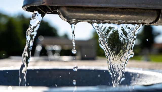 Sunlit stainless steel fountain spout pouring clear water streams, droplets, park close-up