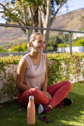 Smiling woman in pink activewear sitting on sunlit balcony turf with bottle and phone