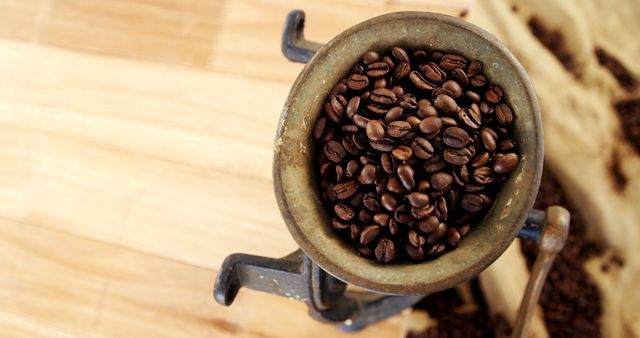 A vintage manual coffee grinder filled with roasted coffee beans sits on a wooden surface, with copy space. Its rustic appearance evokes a sense of traditional coffee preparation methods.