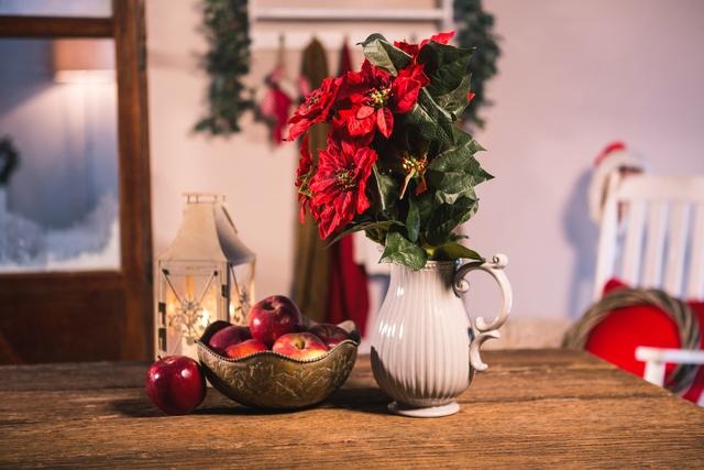 Poinsettia in white vase and bowl of apples on wooden table create a festive and cozy atmosphere. Ideal for holiday season promotions, home decor inspiration, and Christmas-themed content.