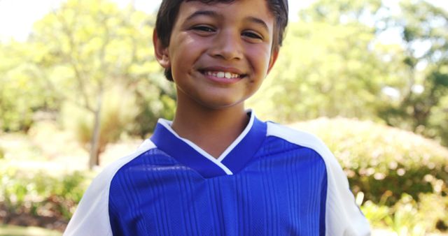 A happy young boy wearing a blue sports jersey smiling outdoors on a sunny day. Background is lush greenery. This image can be used for content related to children, sports, summer activities, outdoor fun, and youth events.