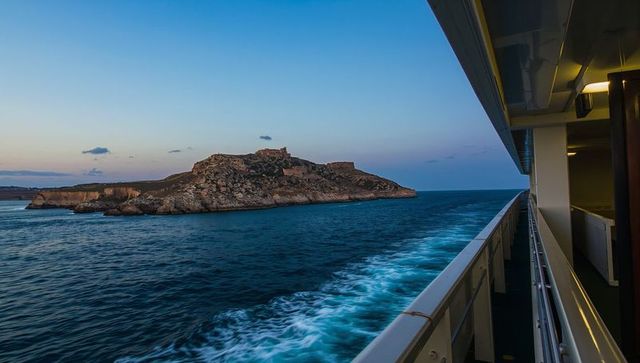 Twilight cruise passing rocky island fortress with foamy wake and open ship balcony view