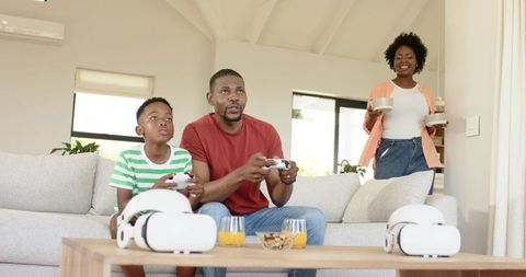 African american dad and son playing video games while mom carrying snacks in modern living room