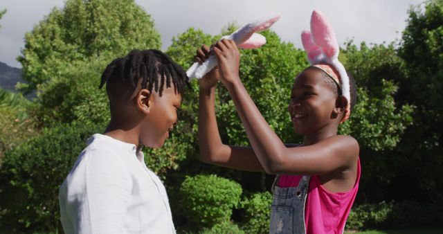 Two children are wearing bunny ears and playing outdoors in a sunny park. They are smiling and enjoying a fun moment together. This image can be used to illustrate themes of childhood joy, outdoor fun, family activities, or the summertime. It is perfect for advertisements, blogs, articles about parenting, family time, or outdoor games.