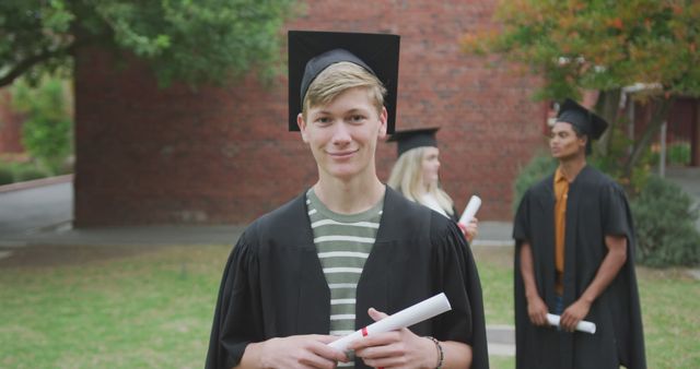Image shows a young male graduate smiling and holding a diploma outdoors. In the background, two other graduates are seen interacting. Perfect for use in educational materials, advertisements for academic institutions, and celebration-themed content emphasizing academic success and achievement.