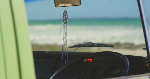 Vintage van dashboard and steering wheel view featuring a colorful dreamcatcher hanging from rearview mirror. Outdoor ocean and beach scenery visible in background, suggesting a summer road trip or adventure. Useful for travel, adventure, and bohemian-themed content.