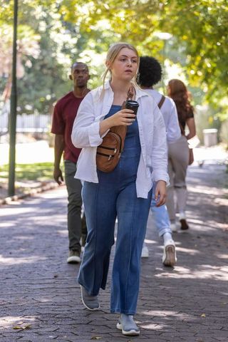 Young woman walking sunlit park holding takeaway coffee wearing denim jumpsuit and crossbody bag