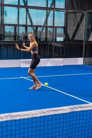 Female athlete playing padel on indoor glass court hitting backhand while wearing smartwatch