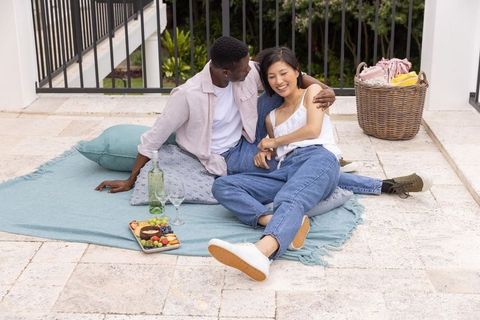Interracial couple enjoying cozy terrace picnic on blue blanket with wine and snacks