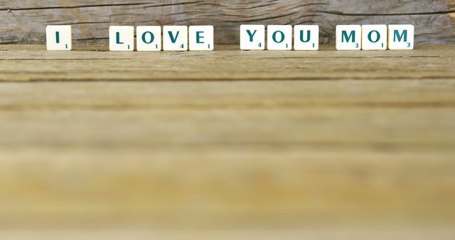 Scrabble tiles spell out 'I Love You Mom' on a rustic wooden table with a wooden background. Useful for Mother's Day cards, social media posts celebrating mothers, or any content related to family affection and appreciation.