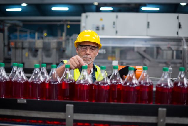 Male factory worker wearing safety helmet and vest is monitoring cold drink bottles on a production line in a drinks production factory. This image can be used to illustrate concepts related to manufacturing, quality control, industrial work, and factory operations. It is suitable for articles, advertisements, and presentations about beverage production, factory safety, and industrial processes.