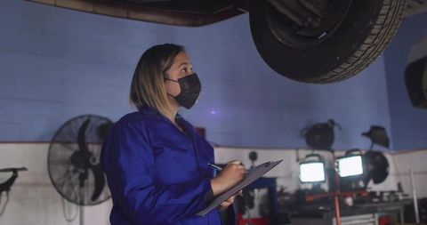 Female technician inspecting undercarriage of lifted car with clipboard and wearing mask in garage