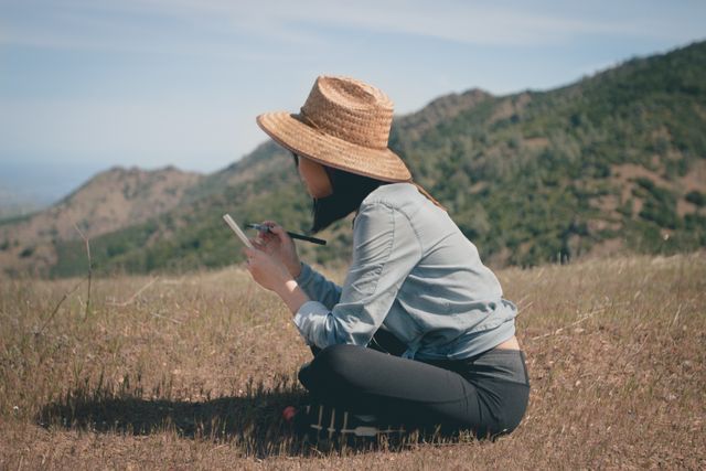 Young woman sitting on the ground while sketching on a notepad in a scenic mountain landscape. Ideal for concepts related to creativity, relaxation, nature, artistic expression, and outdoor activities.