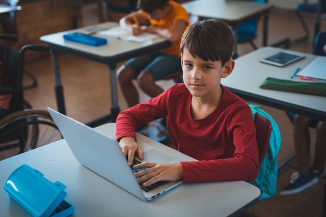 Portrait of caucasian schoolboy sitting at desk in classroom using laptop. childhood, technology and education at elementary school.