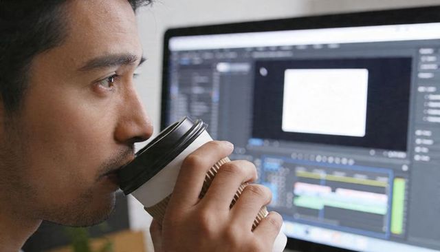 Freelance video editor drinking coffee while focusing on timeline editing at desktop monitor