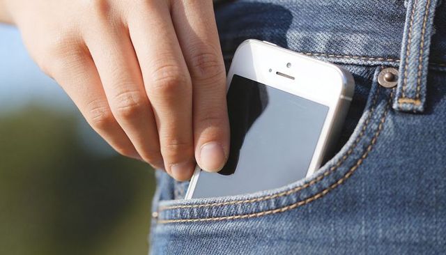 Hand sliding white smartphone into denim jeans front pocket in outdoor casual sunlight