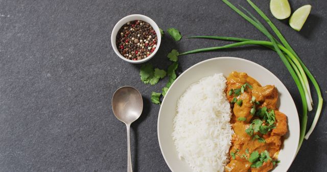 Plate of Indian butter chicken with steamed white rice garnished with cilantro, accompanied by fresh herbs, lime wedges, and a bowl of peppercorns on a gray tabletop. Ideal for use in cooking blogs, recipe websites, culinary magazines, and food presentation showcases.