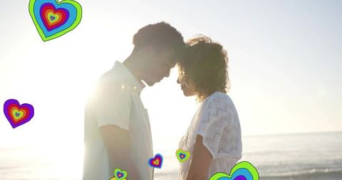 Backlit couple touching foreheads on beach at sunset with colorful heart overlays