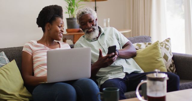 Happy african american couple sitting on sofa talking using laptop and smartphone. staying at home in isolation during quarantine lockdown.