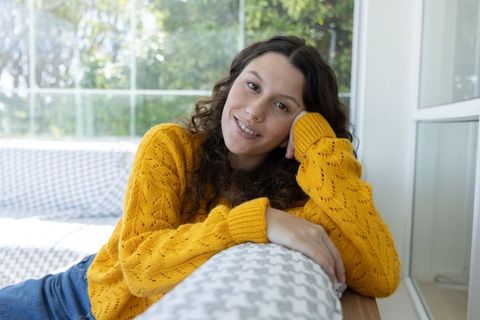 Smiling woman reclining on patterned sofa wearing mustard knit sweater by sunlit window